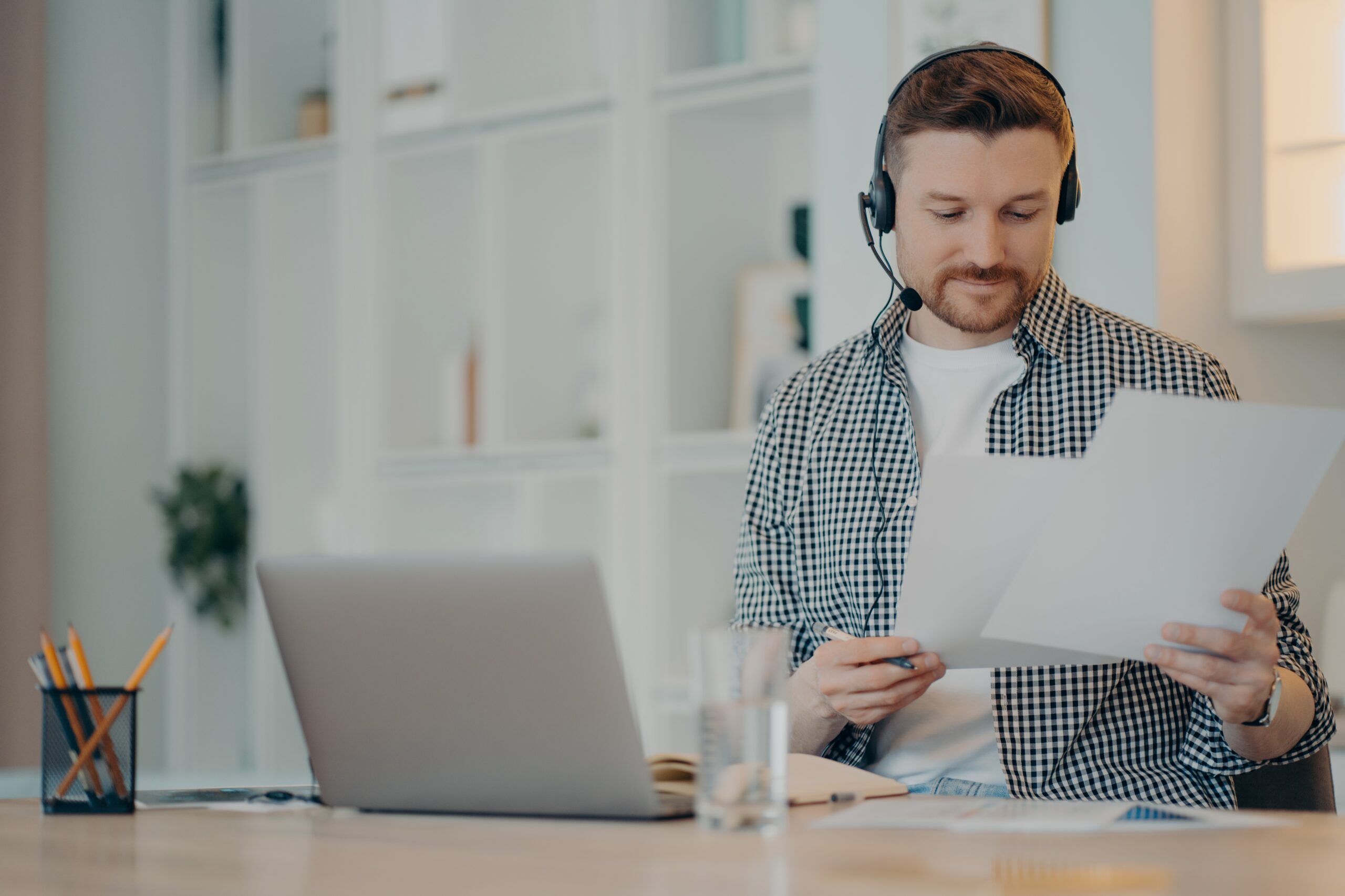 Busy male freelancer wearing headset holding documents in hands and working remotely on laptop while sitting in living room at home. Business online and freelance concept