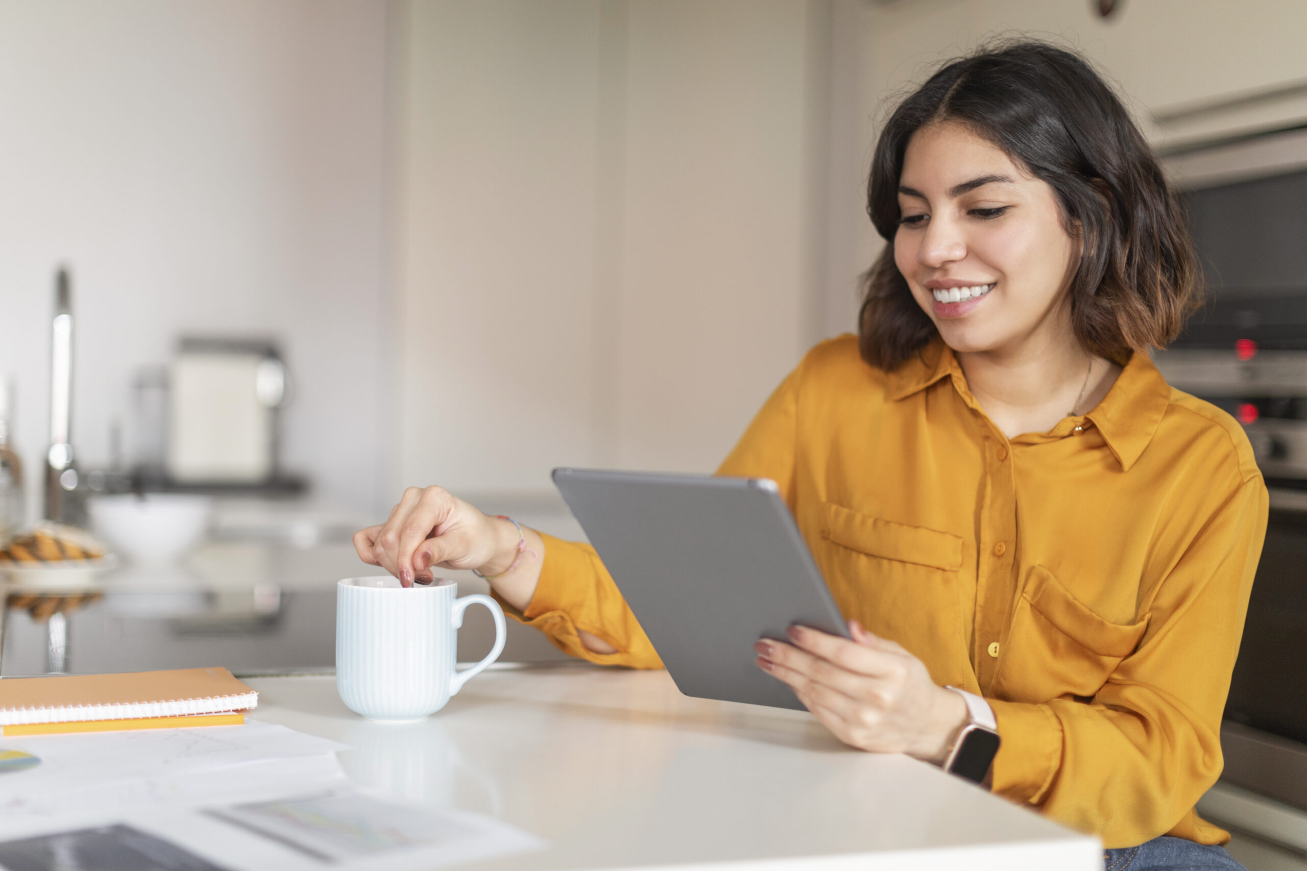Happy Young Arab Woman Relaxing With Digital Tablet In Kitchen And Drinking CoffeeAt Home, Smiling Middle Eastern Female Browsing Internet On Modern Gadget And Enjoying Hot Drink, Free Space